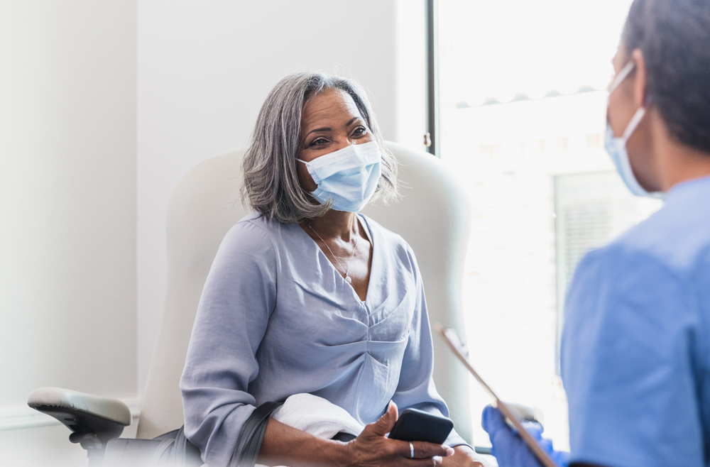 A woman wearing a mask meets with a doctor in a patient room.